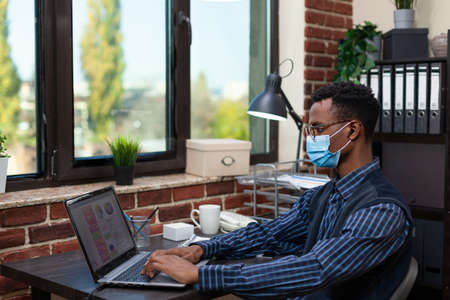 Startup entrepreneur wearing true wireless earbuds and  protective face mask typing sales data on laptop keyboard. Office employee with glasses sitting at desk doing marketing research.の写真素材