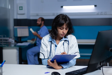 Professional medic looking at computer screen and medical files for healthcare. Doctor analyzing information on monitor and taking notes on papers for assistance, doing overtime work.の写真素材