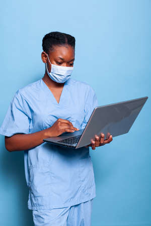 African american therapist nurse with protective face mask to prevent infection with virus typing healthcare treatment on laptop computer in studio with blue background. Medicine serviceの写真素材