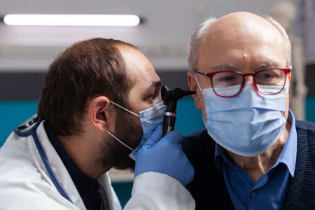 Medic using otoscope to do ear checkup on senior man, doing otolaryngology consultation with patient. Doctor with face mask examining infection to give prescription treatment. Close upの写真素材