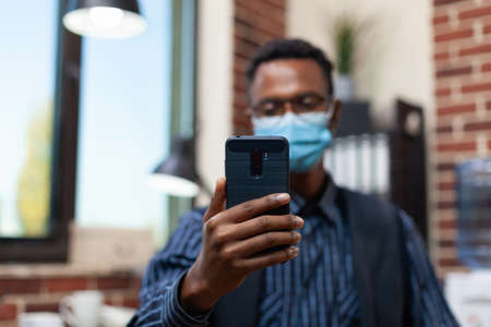 Closeup of startup owner holding smartphone in video call conference wearing protection mask. Focus on employee smart phone in online interview using wireless earbuds.の写真素材