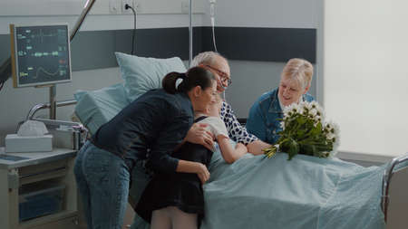 Retired man receiving visit from child and woman with flowers in hospital ward bed. Mother with kid...