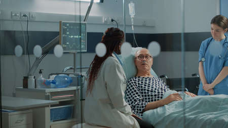Nurse and african american doctor tending to ill patient in hospital ward bed at medical facility. Sick old man with nasal oxygen tube and IV drip bag recovering from injury and diseaseの写真素材