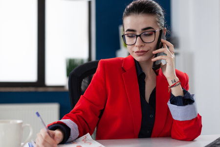 Confident employee with glasses taking notes in phonecall sitting at desk. Small business owner in red jacket writing on paper in startup office. Entrepreneur taking notes in smartphone conversation.の写真素材