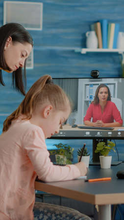 Pupil taking notes on textbook using video call with teacher while mother assisting online class...