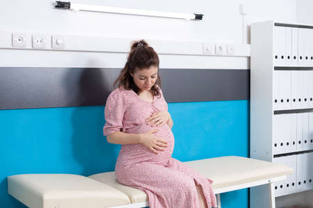 Portrait of woman with belly bump expecting child and sitting in medical office. Patient with...