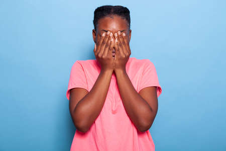 Portrait of scared stressed african american young woman covering face with her hands during photo...