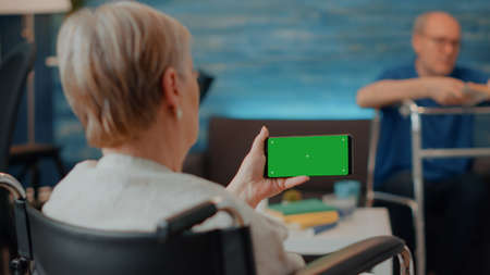 Retiree with disability using horizontal green screen on mobile phone. Elder woman in wheelchair looking at blank copy space with isolated chroma key and mock up template on smartphone.の写真素材