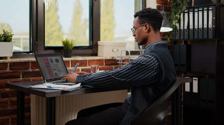 African american person working on e commerce statistics in company office. Business man using laptop to plan commercial report and financial investment for manager career development.の写真素材