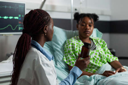 African american practitioner doctor standing beside sick patient holding flu pills bottle explaining healthcare treatment during medical examination in hospital ward. Medicine servicesの写真素材