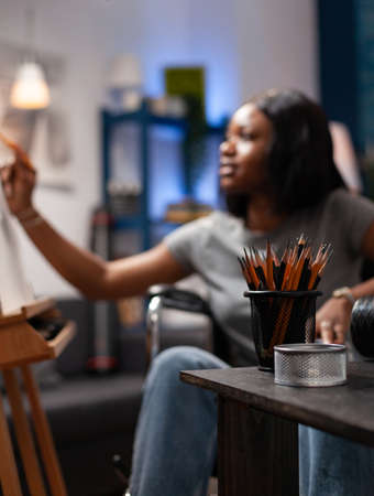 Selective focus on professional support with sketching pencils standing on table in art atelier. In background young artist in wheelchair drawing graphic draw on canvas working at creative masterpieceの写真素材