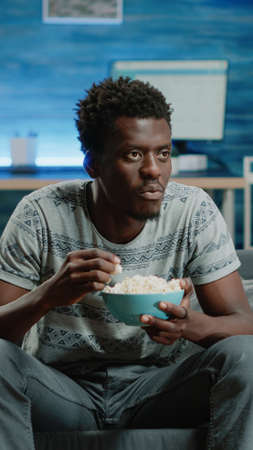 African american man enjoying comedy on television while eating popcorn at home. Black person laughing and watching movie on TV with snack in bowl. Afro adult relaxing on couchの写真素材