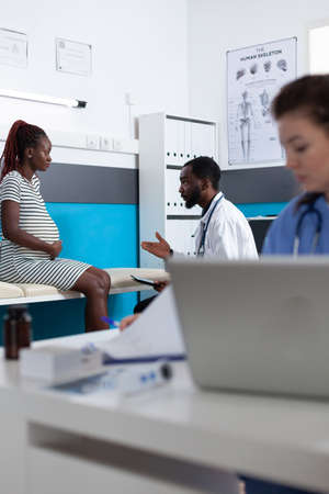 Medic with digital tablet consulting pregnant patient in medical office. Specialist talking to woman expecting baby, receiving healthcare consultation and preparing to give birth.の写真素材