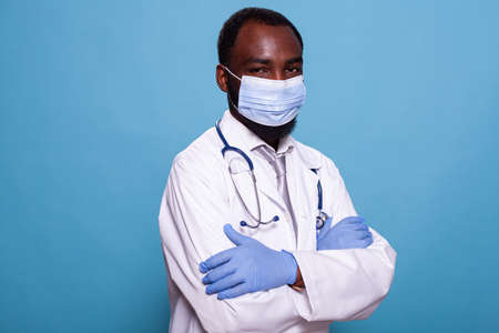 Professional portrait of physician in hospital uniform wearing laboratory coat and protective face mask with arms crossed. African american doctor with stethoscope and medical protective gear.の写真素材