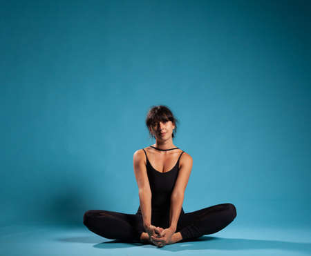 Personal trainer sitting in lotus position on yoga mat stretching body muscles in studio with blue background. Athlete woman wearing sportswear practicing padmasana posture for healthy lifestyleの写真素材