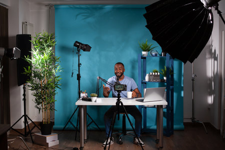 Wide view of smiling influencer interacting with audience sitting at desk holding cup in vlogging...