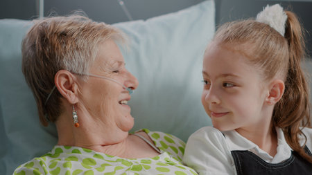 Close up of senior woman with illness enjoying visit from litte girl in hospital ward bed. Aged...
