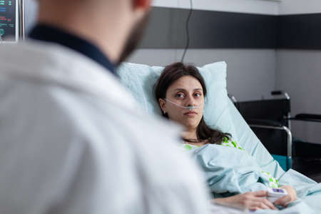 Woman recovering after surgery on hospital bed looking optimistic at doctor explaining treatment options in private ward. Patient with pulse oximeter measuring spo2 talking with medic about illness.の写真素材