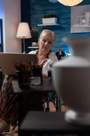 Elderly woman artist living with disability using laptop looking at vase for inspiration in home art studio. Older person sitting in wheelchair holding portable computer browsing sketch ideas.の写真素材