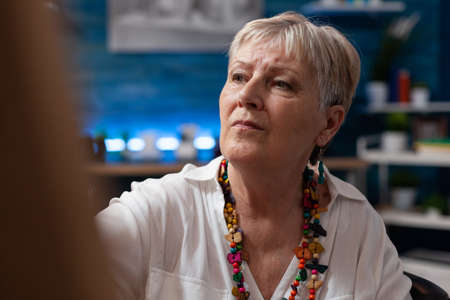 Portrait of gray haired senior lady wearing colorful necklace drawing on canvas in home art studio. Elderly woman sketch artist in front of easel working on creative freehand charcoal painting.の写真素材
