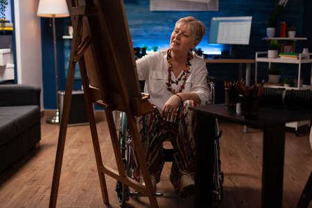 Elderly woman sitting in wheelchair drawing black and white sketch using pencil on canvas in artist...