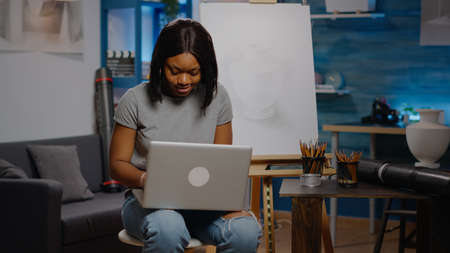 Black young artist typing on laptop for art project while sitting in workshop space at home. African...