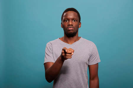 Portrait of young man pointing index finger at camera, standing over blue background. Focused person indicating direction and making choice to choose someone in studio. Individuality pickの写真素材
