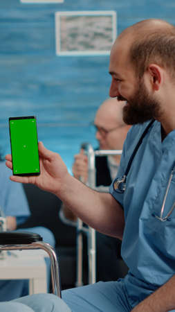 Man nurse holding green screen on smartphone in nursing facility. Old woman sitting in wheelchair and medical assistant looking at chroma key with mockup template and isolated backgroundの写真素材