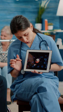 Patient with disability looking at virus animation on digital tablet and nurse explaining coronavirus pandemic and bacteria. Specialist talking to senior man sitting in wheelchairの写真素材