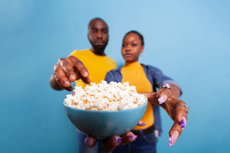 Man and woman holding glass bowl with popcorn and eating snack while they watch movie on television. Cheerful couple enjoying film on tv with food, smiling together in front of camera.の写真素材