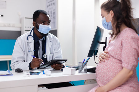 Pregnant adult meeting with doctor at checkup appointment during coronavirus pandemic. Patient with pregnancy talking to practitioner about birth and medical care, wearing face masks.の写真素材