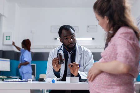 African american medic giving bottle of pills to pregnant person to cure illness. Physician holding flask with prescription medicine and treatment to give to pregnant woman.の写真素材