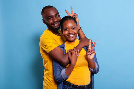 Couple hugging and showing peace sign with fingers on camera, feeling happy in relationship. Portrait of joyful man and woman in embrace doing v symbol gesture with hands and smiling.の写真素材