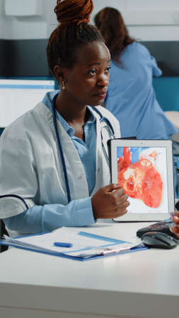 Doctor showing tablet with heart figure for cardiology checkup to patient at consultation. Young parent looking at device with organ for cardiovascular and blood flow healthcare.の写真素材