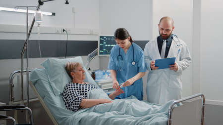 Medical team doing checkup visit for aged patient with disease in hospital ward bed. Doctor and nurse consulting senior woman with oximeter for oxygen saturation and IV drip bag.の写真素材