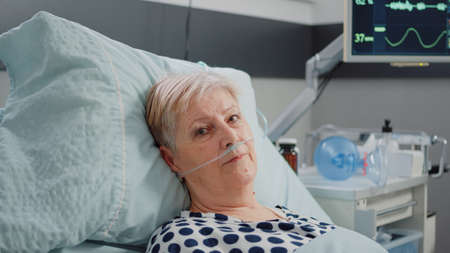 Portrait of elder patient with oxygen tube laying in hospital ward bed. Woman with illness looking at camera while waiting for healthcare assistance in emergency room for intensive careの写真素材