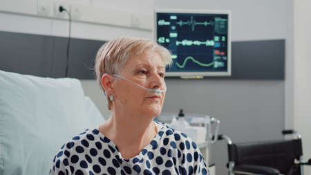 Close up of retired woman with nasal oxygen tube waiting on assistance. Portrait of person with respiratory disease having heart rate monitor for pulse measurement in hospital wardの写真素材