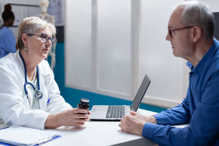Woman medic giving bottle of pills to aged man with illness at checkup visit. General practitioner holding jar of prescription medicine for treatment against diagnosis. Medical appointmentの写真素材