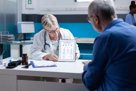Physician holding tablet with image of human skeleton to explain diagnosis to old man in cabinet. Doctor and patient looking at bones and spinal cord illustration on device for examination.の写真素材
