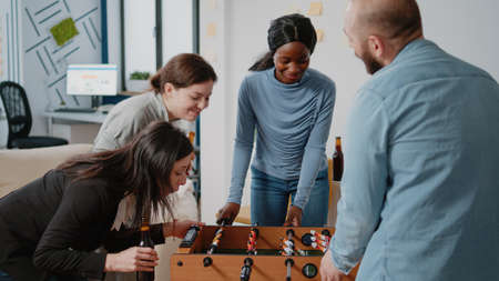 Diverse group of people enjoying fun activity after work with foosball game table and drinks. Workmates playing soccer game, drinking beer and eating pizza while celebrating after hoursの写真素材