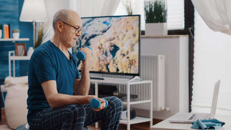 Man lifting dumbbells while sitting on fitness toning ball an looking at online video of workout on laptop. Retired adult doing exercise with weights and watching training lesson on deviceの写真素材