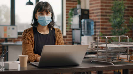 Portrait of employee with face mask using laptop to work on business strategy and project planning...