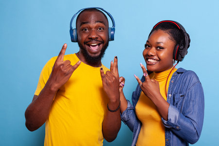 Happy people showing rock symbol with fingers on camera, listening to music on headphones. Playful couple enjoying song and rhythm on headset, doing hands gesture and smiling.の写真素材