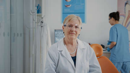 Portrait of mature stomatologist looking at camera in cabinet, working with dental tools for oral care. Woman orthodontist wearing protective glasses to treat dentition problemsの写真素材
