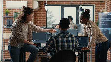 System engineer sitting on desk discussing with mixed team of coders about ai innovation in front of multiple screens compiling code. Programmers doing teamwork looking at machine learning algorithms.の写真素材