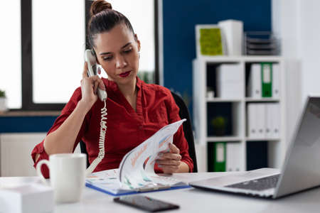 Entrepreneur in red shirt analyzing chart in phonecall. Businesswoman sitting at desk looking at...