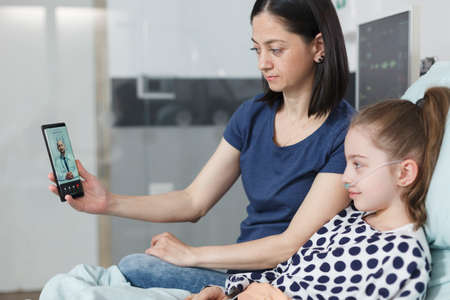 Young mother and sick little child talking with pediatrician on smartphone videocall. Woman and ill girl talking with general practitioner on teleconference while using phone conference app.の写真素材