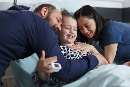 Happy young parents hugging hospitalized recovered little daughter while in pediatric clinic patient room. Cheerful family laughing and hugging in health pediatric hospital room.の写真素材