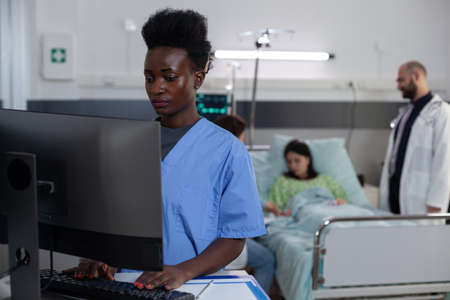 African american nurse using personal computer for clinical imaging in patient private hospital ward. Medical doctor and physician consulting patient while attendant is reading lab results.の写真素材