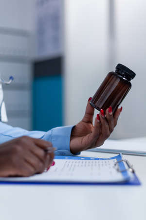 Close up of hospital general practitioner specialist holding a vial of medicine while checking symptom list and patient illness history. Medic having antiviral bottle in handの写真素材